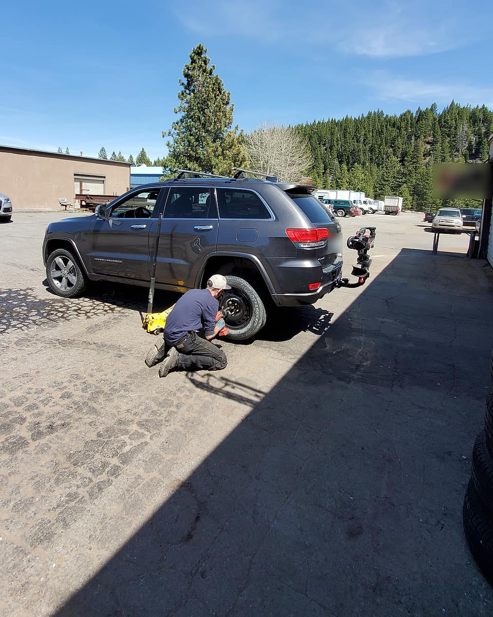 A tech in a cap kneeling at the rear wheel of a gray Jeep with a yellow floor jack, pines and blue sky behind