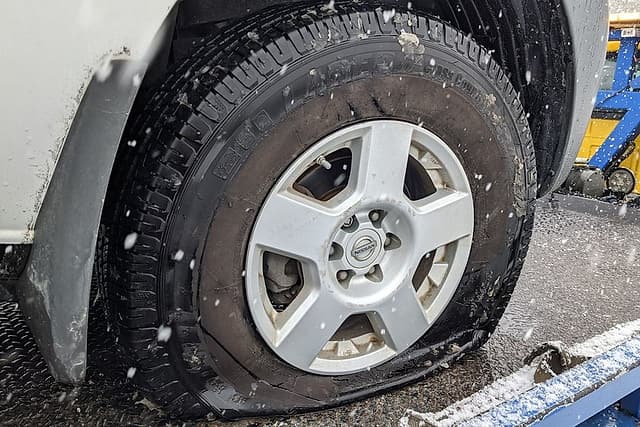 A flat tire on a silver Nissan SUV riding a flatbed tow truck in falling snow
